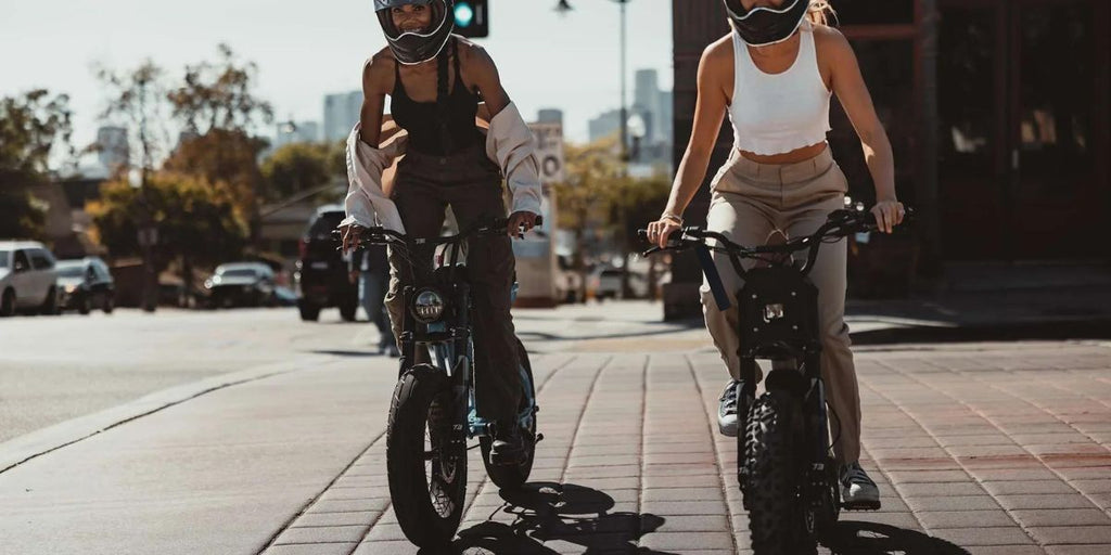 Two ladies in black and white outfits with helmets riding fat-tire e-bikes in the city.