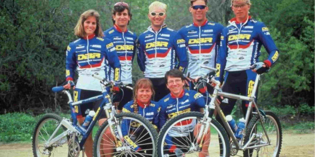 Seven DBR players in blue uniforms posing in front of woods with two DBR bikes placed in front of them.