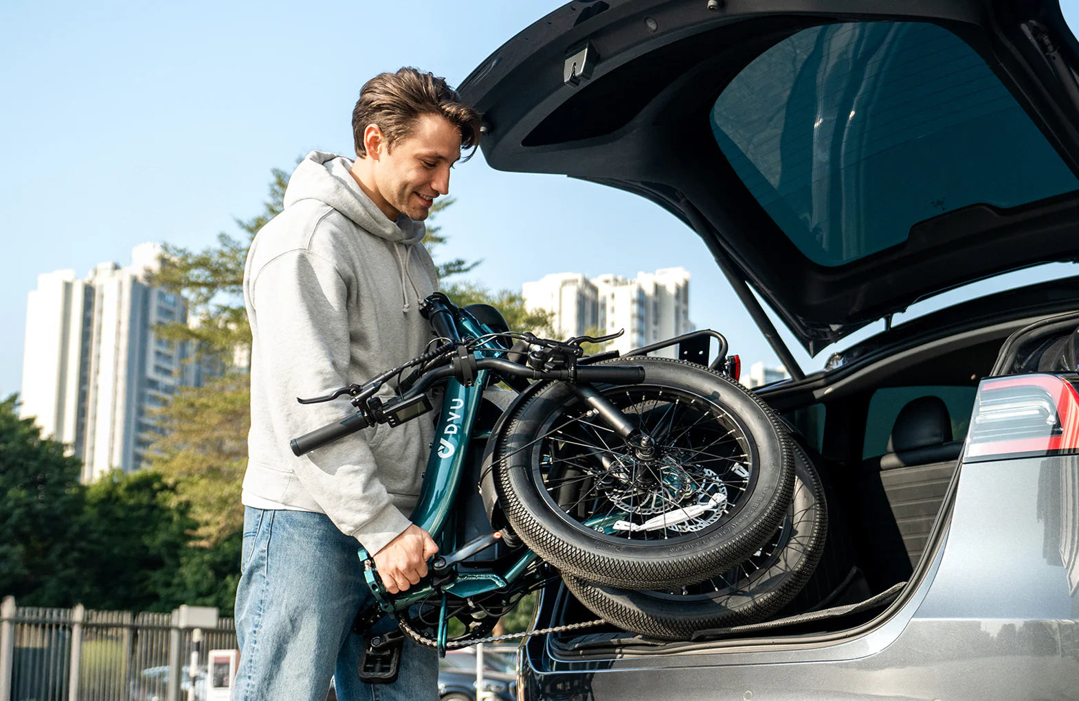 Man loading a folded DYU C9 electric bike into the trunk of a car with cityscape in the background