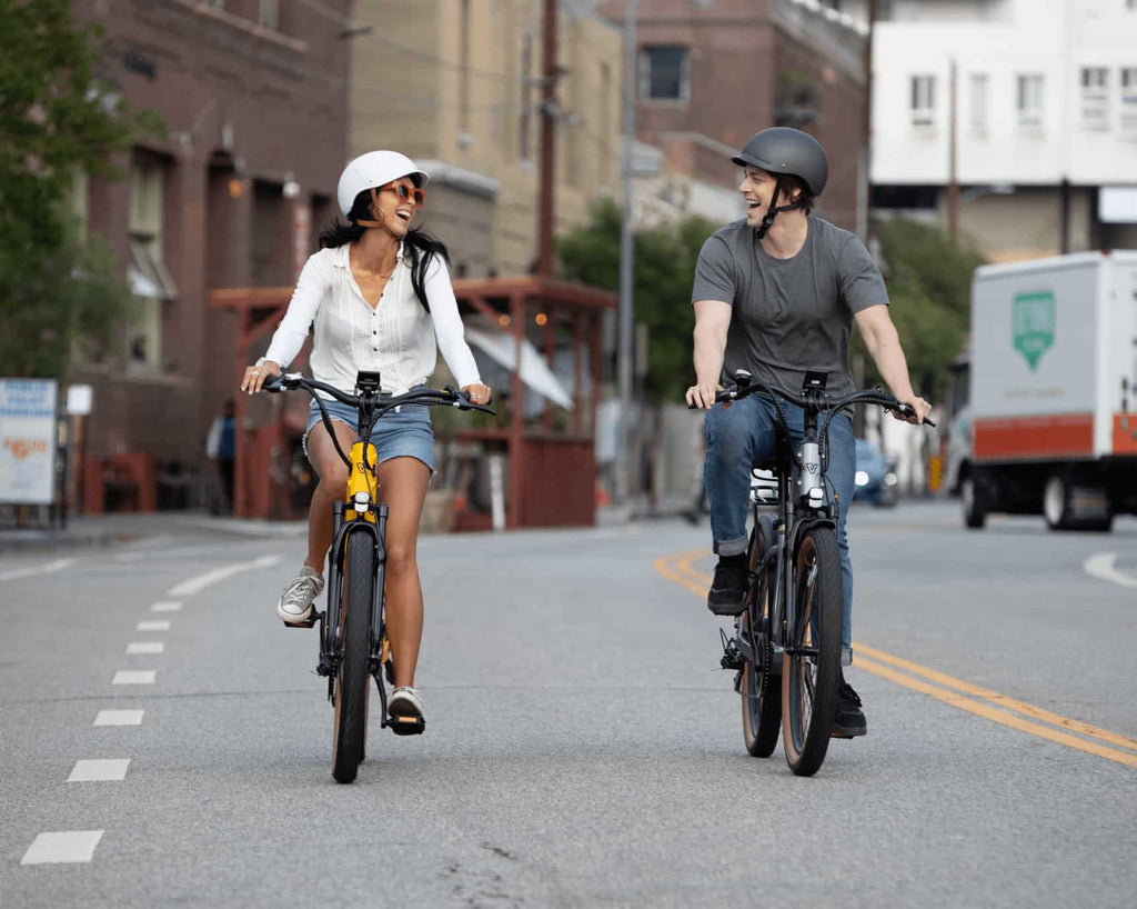 A man and woman happily ride electric bikes on a highway, with smiling faces and scenic background.