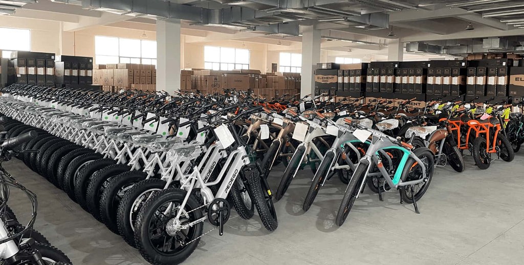 Organized warehouse at Damaxed e-bike factory with silver, orange, and green assembled bikes, neatly surrounded by packaging boxes ready for shipment.