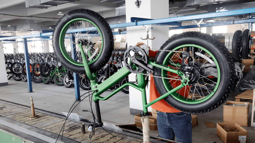 Orange-clad engineer assembling green e-bike on production line with rows of cars awaiting quality inspection in background.