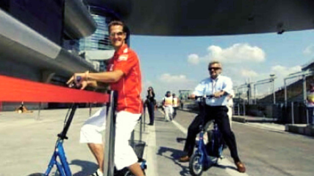Two F1 staff members, wearing red shirts, ride DBR-sponsored electric scooters in F1 arena event.