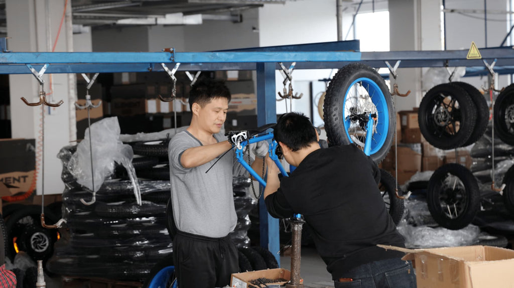 Two engineers assembling blue electric bike on factory production line with neatly arranged tires in background.