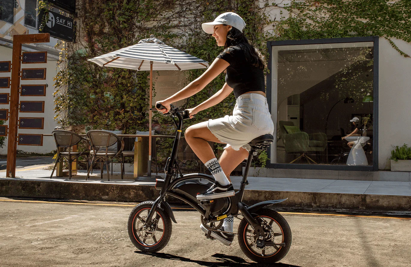 Person riding a small dyu d3f foldable electric bike on a sidewalk with outdoor seating in the background