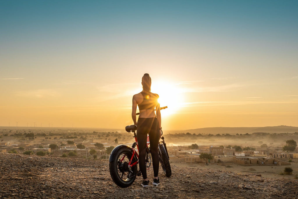 A woman in a sports wear standing in front of a DAMAXED electric fat-tire bike during a desert sunset.