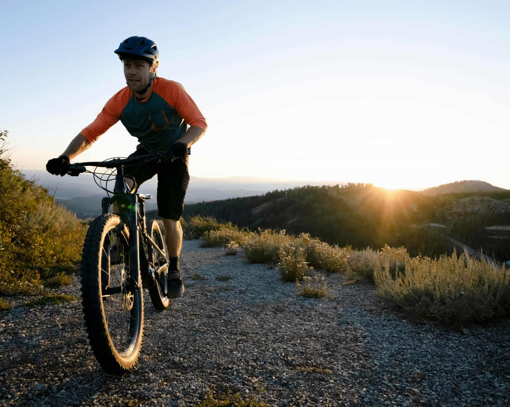 A man in cycling clothes and helmet rides DAMAXED electric mountain bike on mountain, with sunset in the background.