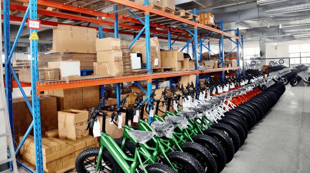 Neatly placed rows of green and orange e-bikes in front of shelves filled with paper boxes. More rows of e-bikes in the distance.