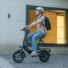 Person riding a C3 foldable electric bike with a helmet and backpack in front of a building.