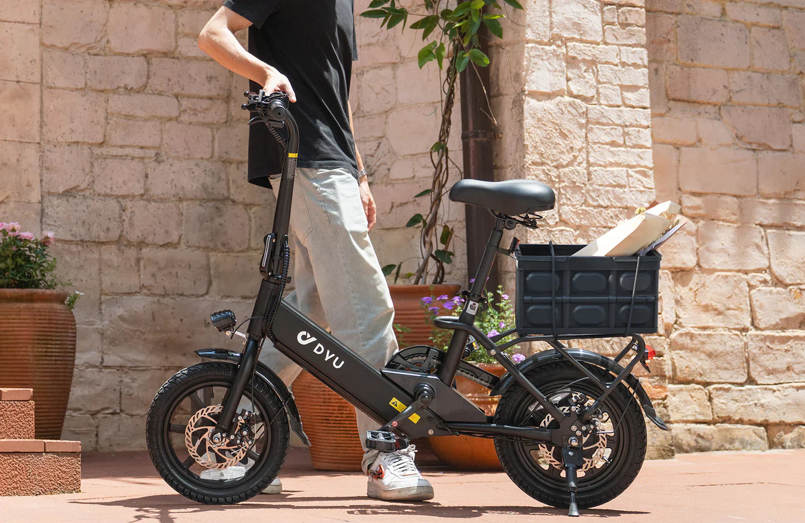 Person holding a black C3 foldable electric bike with a basket against a stone wall background