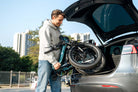 Man loading a folded DYU C9 electric bike into the trunk of a car with cityscape in the background