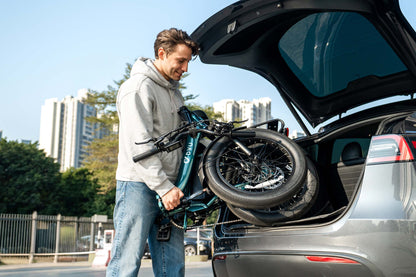 Man loading a folded DYU C9 electric bike into the trunk of a car with cityscape in the background
