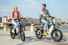 Two people riding DYU C9 electric bikes by a waterfront with buildings in the background