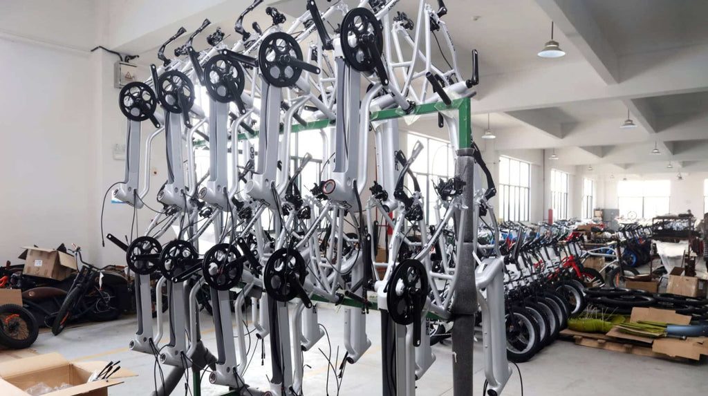 Silver frame neatly displayed for assembly, with rows of assembled e-bikes in background awaiting quality inspection.